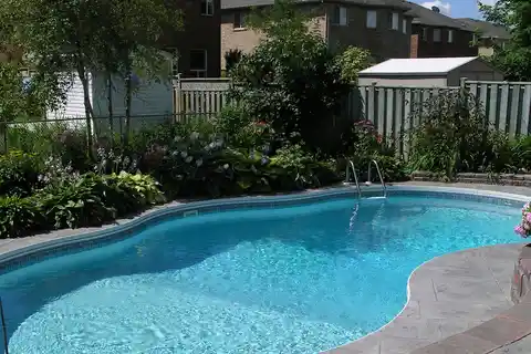 A fenced backyard inground pool with a stone patio and landscaping
